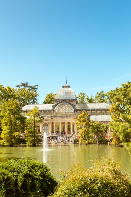 A historic glass conservatory with a domed roof and ornate architectural details, situated amidst lush green trees around a reflective pond. The bright blue sky overhead highlights the sunlight illumi