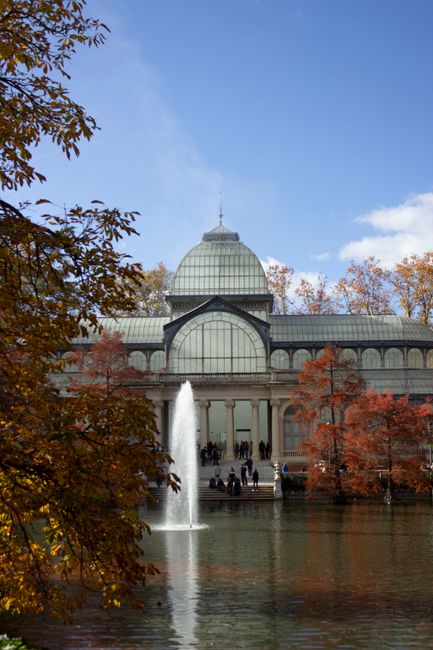 The image shows a large, elegant conservatory with a domed glass roof and arched windows, situated behind a pond with a central fountain. Surrounding the pond are trees with autumn foliage in shades o