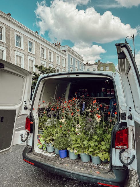 The back of a white delivery van open to reveal a variety of fresh flowers in different stages of bloom, including bright red poppies, white daisies, and lush green foliage, arranged in small blue and