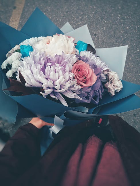 A floral gift bouquet featuring a large white chrysanthemum, a pale pink rose, and smaller purple and white flowers, arranged with green foliage. The bouquet is wrapped in blue and white paper with a 