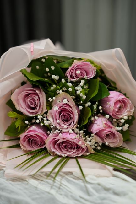 A bouquet of fresh, pastel pink roses with tightly layered petals, accented by small white baby's breath flowers and lush green leaves. The arrangement is wrapped in white tissue paper with a subtle p