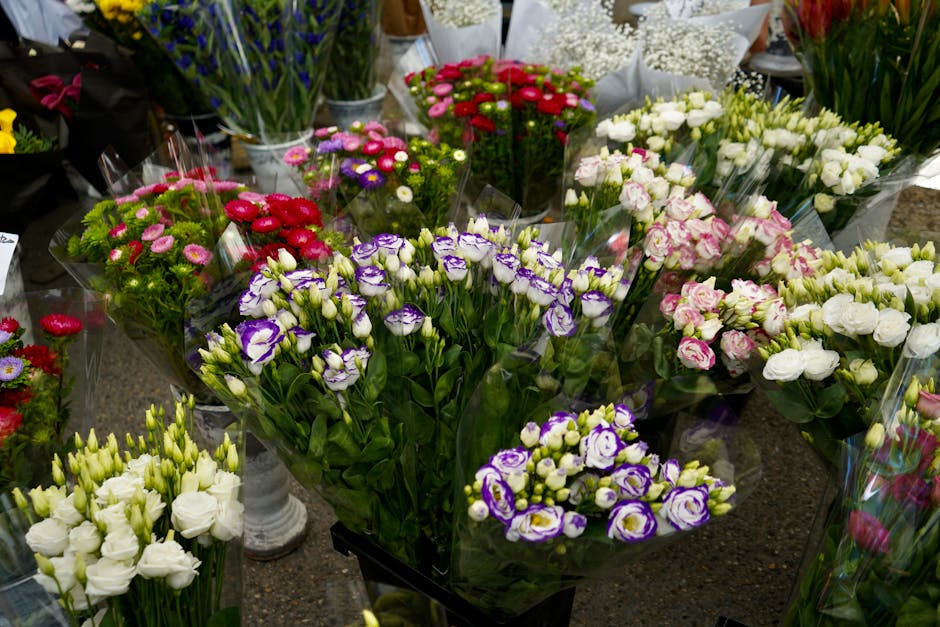 A vibrant array of fresh flowers arranged in bouquets at a flower shop, featuring purple and white irises, white roses, pink and white lisianthus, red and pink daisies, and various other blooms. The b